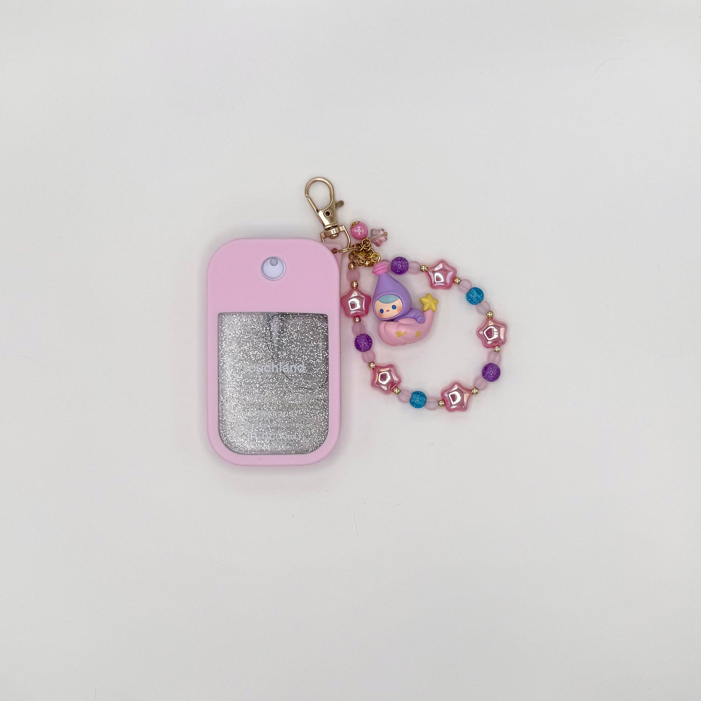 Touchland Power Mist hand sanitizer in a pink silicone case with a glitter window, styled on a lavender bow bag. Attached charm features a Pucky chibi figure in a purple flower hood with blue accents, dangling from a beaded strand of purple glitter, lavender, and gold beads with a pink ball accent. TOUCHLAND NOT INCLUDED WITH THIS PRODUCT!