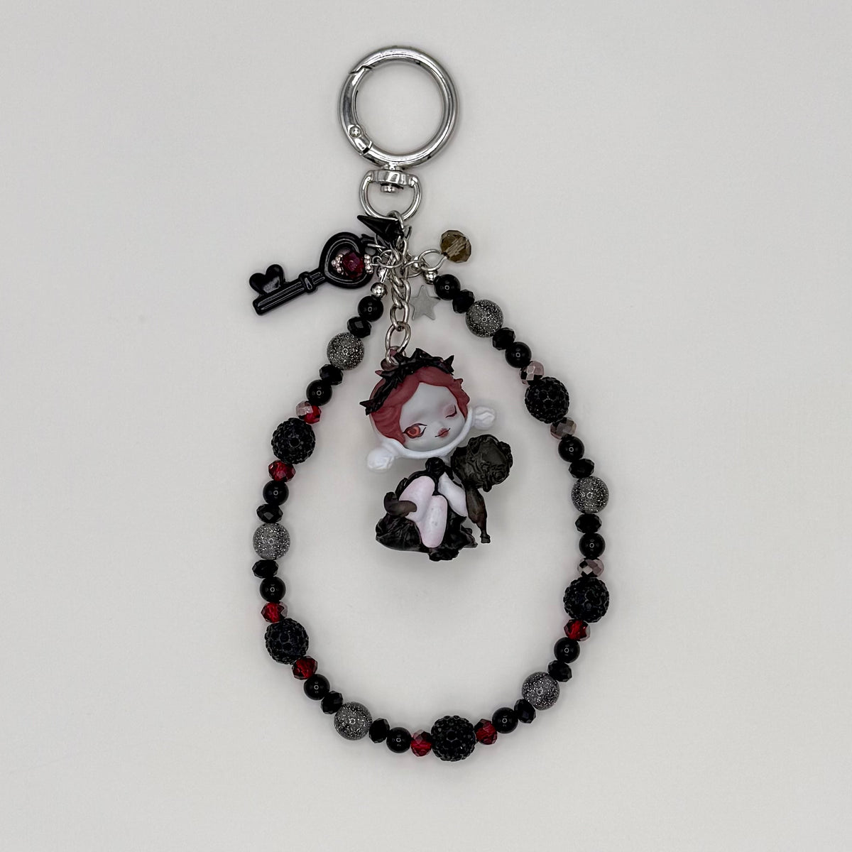 Beaded keychain with black faceted crystal, dark red crystal, and silver glitter beads with silver spacers, featuring a red-haired character figure in a black outfit hugging a dark companion figure, with a black heart key charm and smoky crystal charm on a silver ring clasp.
