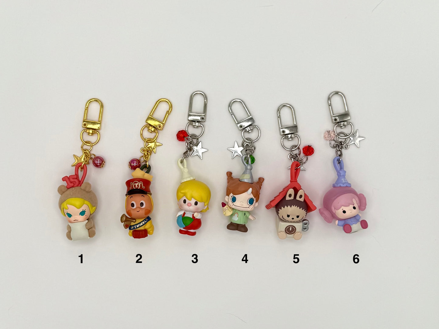 Six numbered character keychains on a white background. Features: (1) blonde girl in a bear hood with gold clasp, bell and star charms, (2) brown character in a red marching band uniform with gold clasp, (3) blonde boy holding a ball with silver clasp and bead charms, (4) freckled girl in brown outfit with silver clasp, (5) hedgehog in a red house hat with clock detail, (6) pink boy in lavender outfit with crystal charms.