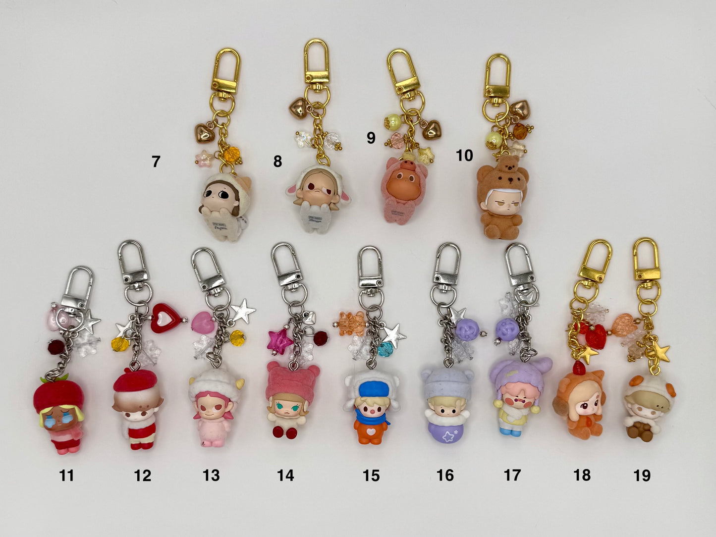 Thirteen numbered flocked-texture character keychains on a white background. Top row features soft round figures: calico cat (7), white sheep (8), pink bear (9), and brown bear (10) with gold clasps and heart charms. Bottom row features chibi dolls in costumes including red tomato hat (11), red beret (12), pink cat ears (13–14), blue & white hat (15), lavender and purple bear ears (16–17), orange fox (18), and small brown sheep (19) with silver and gold clasps.