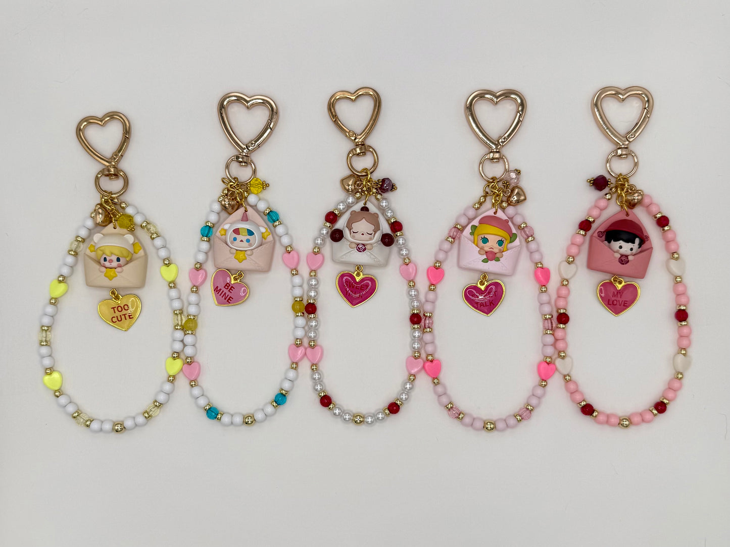 Five kawaii phone charm keychains displayed in a row on a white background, each featuring a different chibi figure peeking out of a pastel envelope with a heart enamel charm and gold heart-shaped clasp. Variants include Sweet Bean, Pucky, SP, Molly, and Haci, each with a unique color-coordinated beaded loop strand.