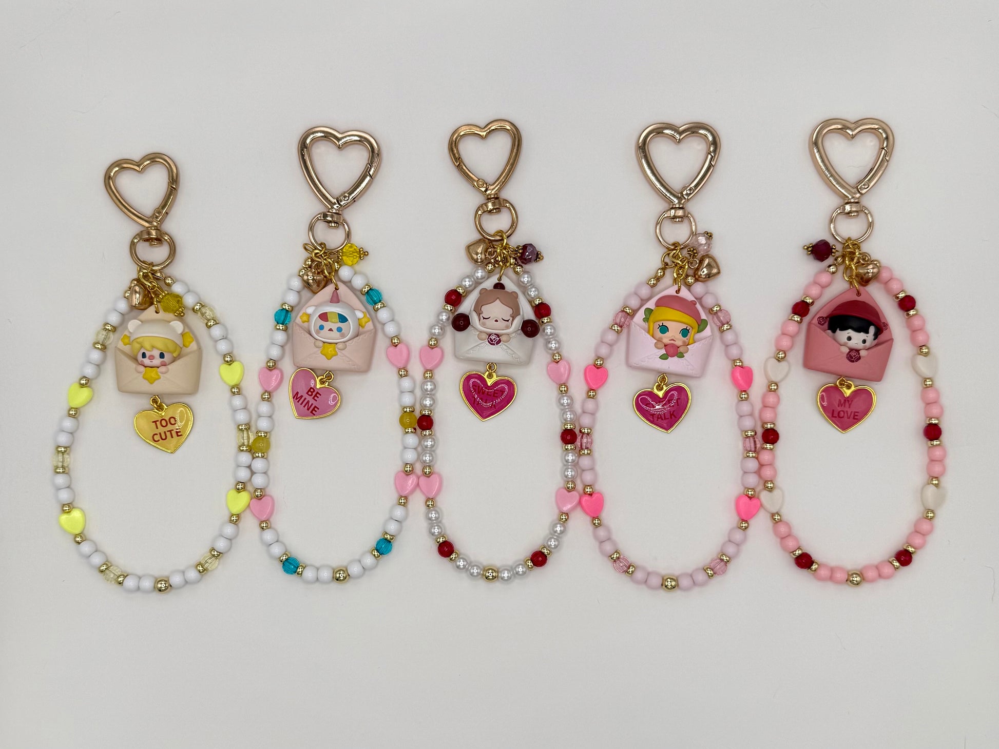 Five kawaii phone charm keychains displayed in a row on a white background, each featuring a different chibi figure peeking out of a pastel envelope with a heart enamel charm and gold heart-shaped clasp. Variants include Sweet Bean, Pucky, SP, Molly, and Haci, each with a unique color-coordinated beaded loop strand.