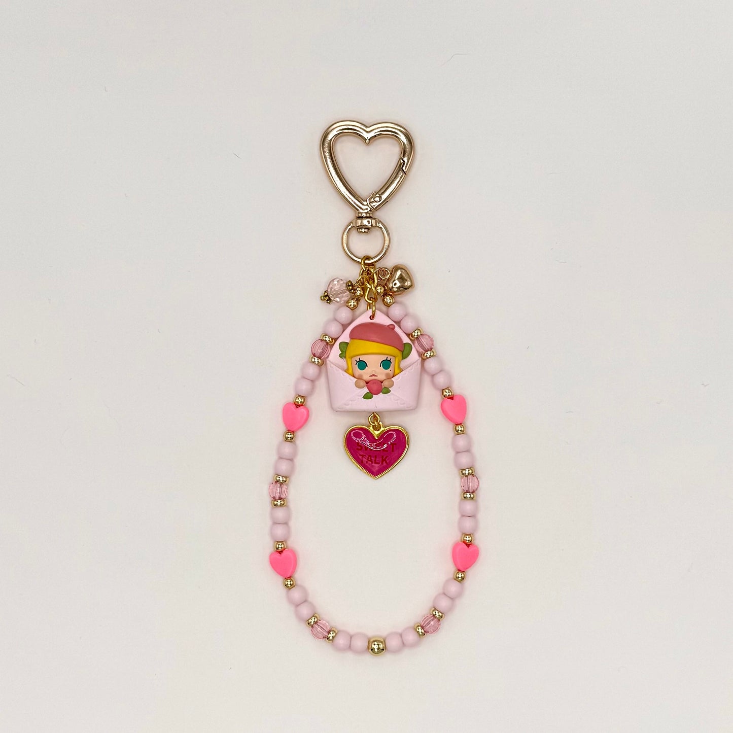 Kawaii phone charm keychain featuring a Molly chibi figure in a yellow hat peeking out of a lavender envelope, with a 'Sweet Talk' hot pink heart enamel charm, silver heart-shaped clasp, and a beaded loop strand of lavender, pink heart, and clear pink beads with gold accents.
