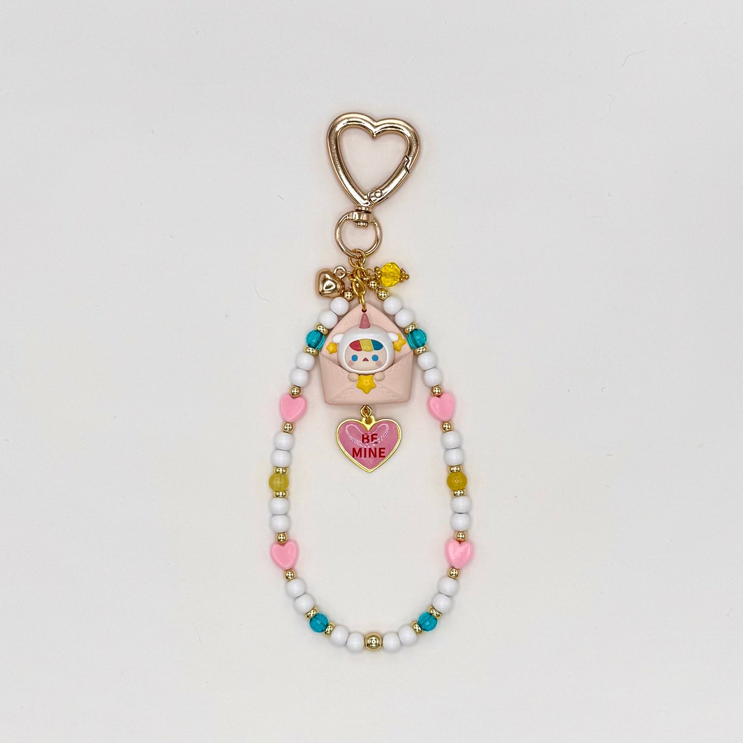 Kawaii phone charm keychain featuring a colorful Pucky chibi figure peeking out of a pink envelope, with a 'Be Mine' pink heart enamel charm, gold heart-shaped clasp, and a beaded loop strand of white, pink heart, teal, and olive beads with gold accents.