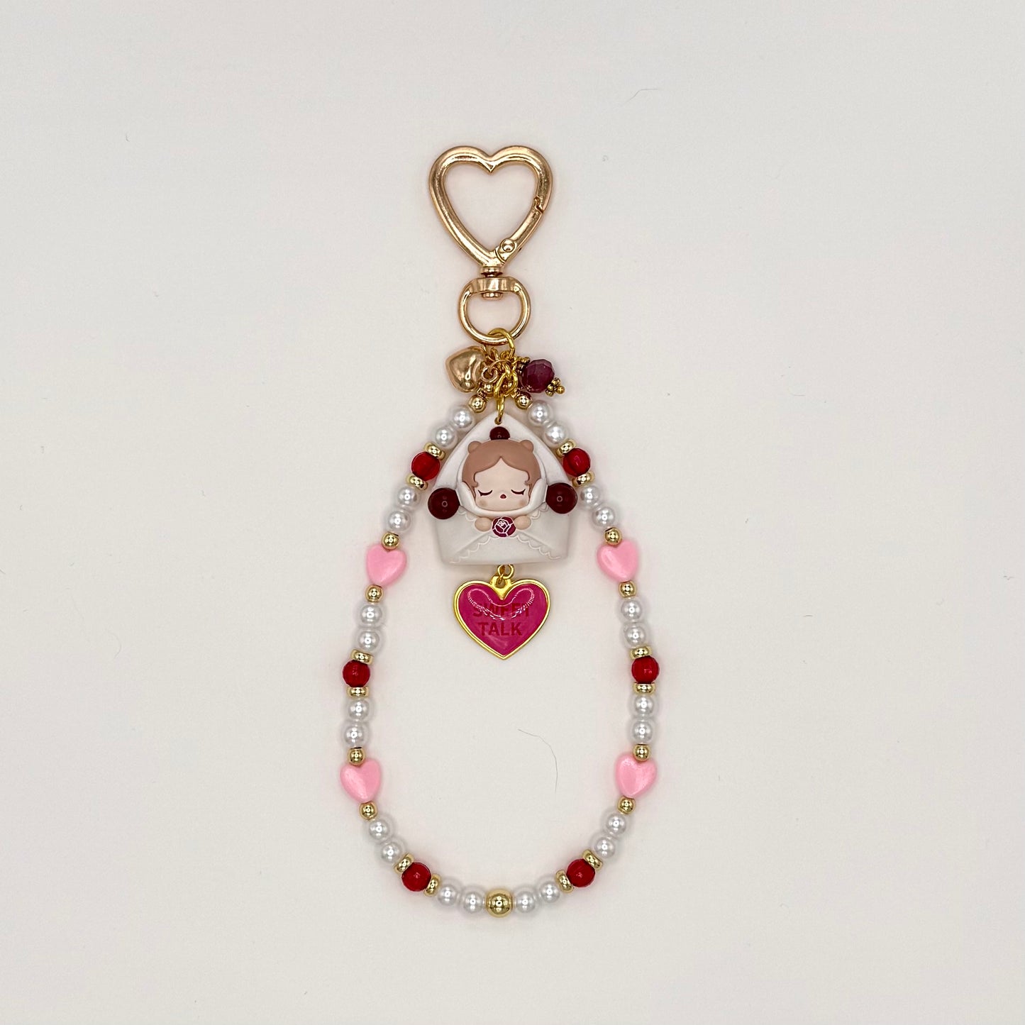 Kawaii phone charm keychain featuring a sleepy chibi girl peeking out of a white envelope, with a 'Sweet Talk' pink heart enamel charm, gold heart-shaped clasp, and a beaded loop strand of white pearls, red, and pink heart beads with gold accents.