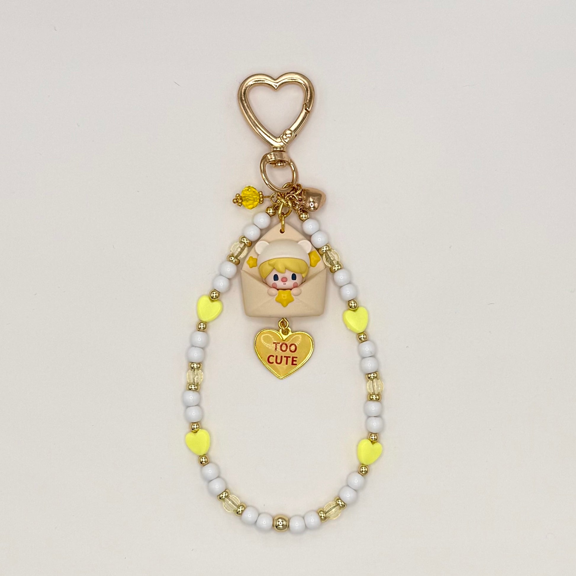 Kawaii phone charm keychain featuring a Sweet Bean chibi figure in a bear hood peeking out of a cream envelope, with a 'Too Cute' gold heart enamel charm, gold heart-shaped clasp, and a beaded loop strand of white, yellow heart, and clear beads with gold accents.