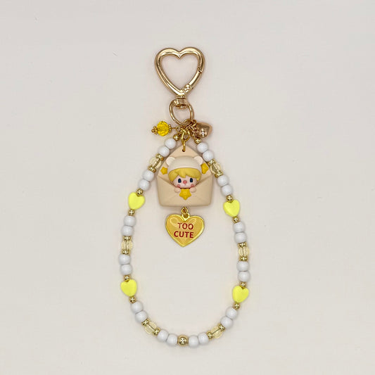 Kawaii phone charm keychain featuring a Sweet Bean chibi figure in a bear hood peeking out of a cream envelope, with a 'Too Cute' gold heart enamel charm, gold heart-shaped clasp, and a beaded loop strand of white, yellow heart, and clear beads with gold accents.