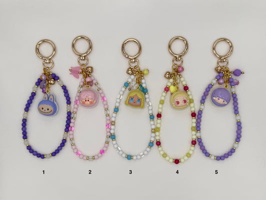 Five numbered kawaii wrist strap keychains on a white background, each with a gold spring ring clasp and a macaron-shaped dome charm containing a chibi character face. Variants include a purple strap with blue bunny charm (1), pink and mauve strap with pink piggy charm (2), white and blue strap with yellow Alice-style charm (3), yellow-green and red strap with pink chibi charm (4), and an all-purple strap with blue character charm (5).