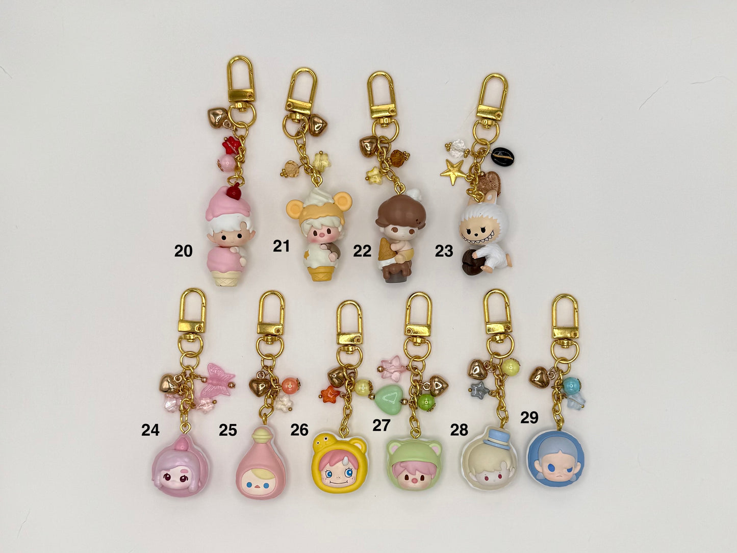 Ten numbered kawaii character keychains with gold clasps on a white background. Includes a pink elf girl (20), cream mouse in ice cream cone costume (21), mushroom-cap boy (22), white wolf with star charms (23), pink ghost figure (24), pink potion bottle (25), yellow bear face (26), green hooded character (27), and two clear dome figures with characters inside (28–29), all with decorative bead and chain accents.