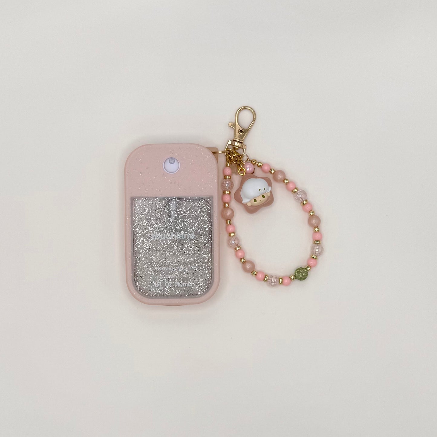 Touchland Power Mist hand sanitizer in a pink silicone case with a glitter window on a white background. Attached gold lobster clasp keychain features a Dimoo chibi charm in a yellow star hat, paired with a beaded wrist strap of pink, orange, and gold star-shaped beads with gold accent spacers. TOUCHLAND NOT INCLUDED WITH THIS PRODUCT!