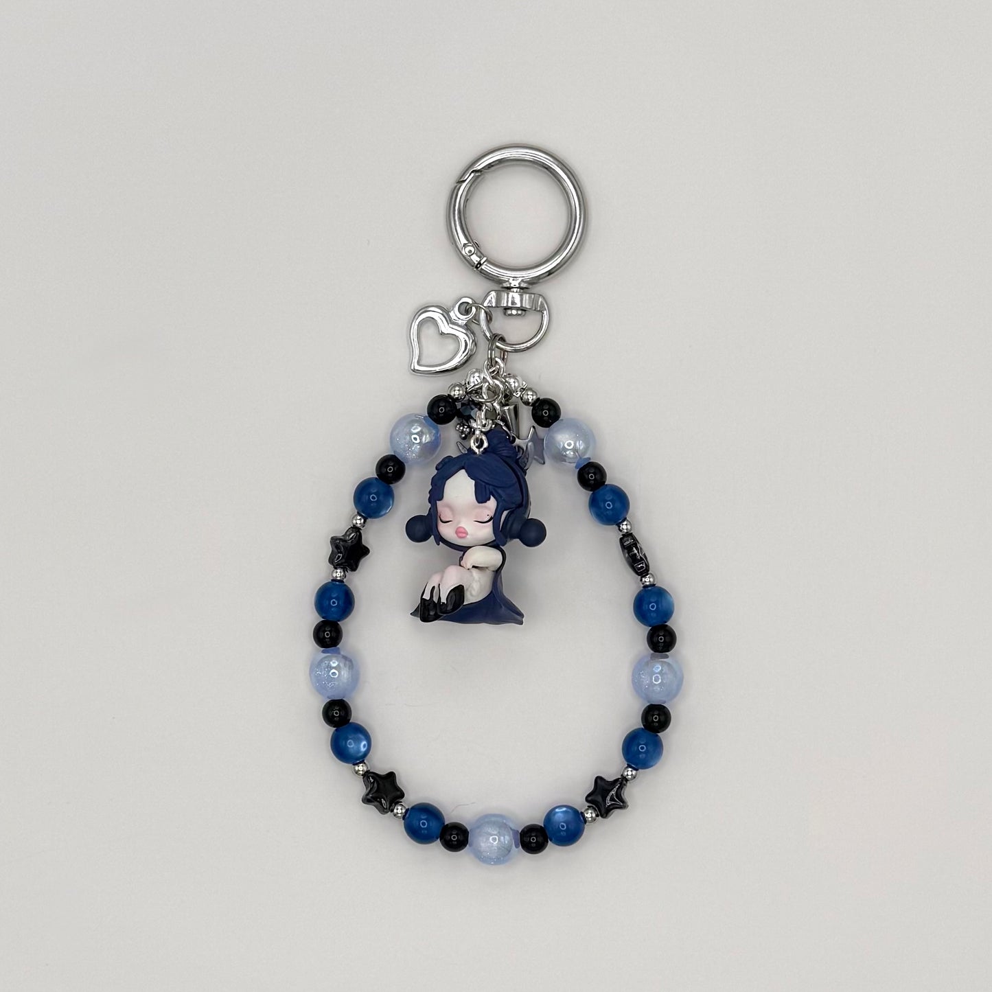 Beaded keychain with deep blue cat eye, pale blue frosted, and black star beads with silver spacers, featuring a dark blue-haired character girl figure in a navy dress, with a silver heart charm and small star charm on a silver ring clasp.
