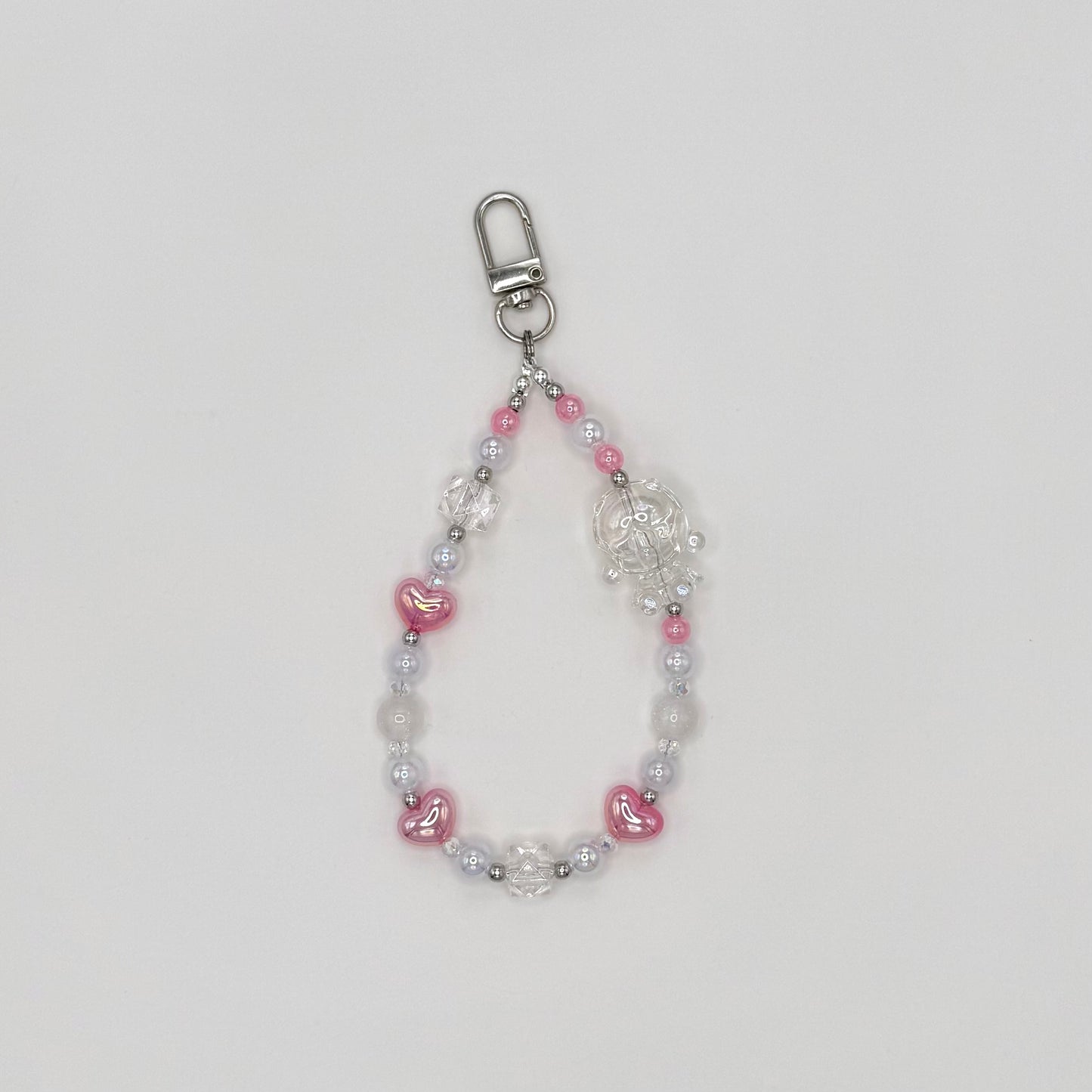Phone wrist strap with pink heart-shaped beads, frosted white round beads, clear crystal bicone beads, and silver spacers, featuring a clear transparent character charm, on a silver swivel clasp."\