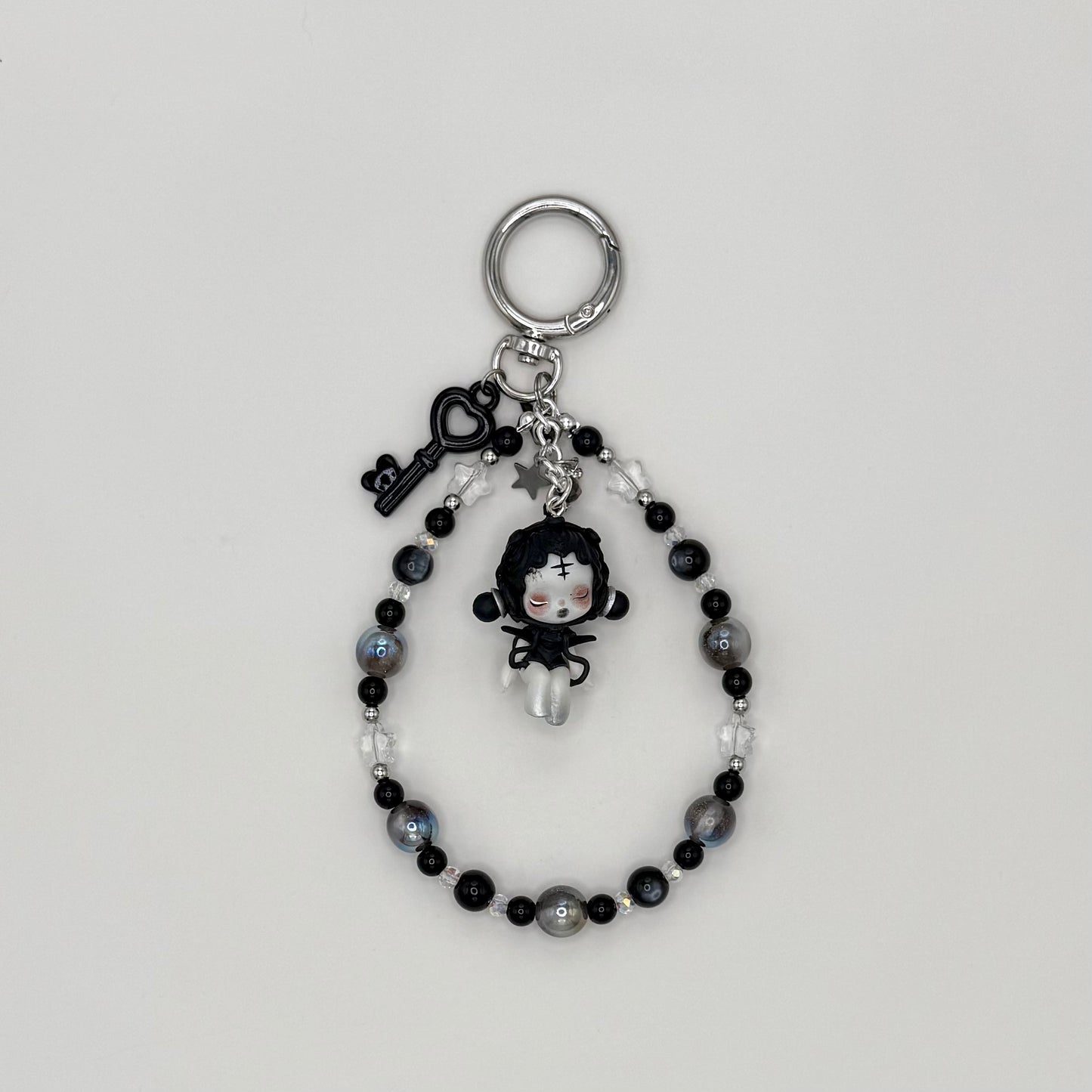 Beaded keychain with gray resin, black cat eye resin, and clear crystal beads with silver spacers, featuring a gothic black-haired character figure in a black dress with a cross on her forehead, with a black heart key charm and silver star charm on a silver ring clasp.