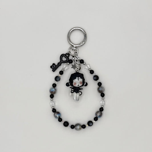 Beaded keychain with gray resin, black cat eye resin, and clear crystal beads with silver spacers, featuring a gothic black-haired character figure in a black dress with a cross on her forehead, with a black heart key charm and silver star charm on a silver ring clasp.