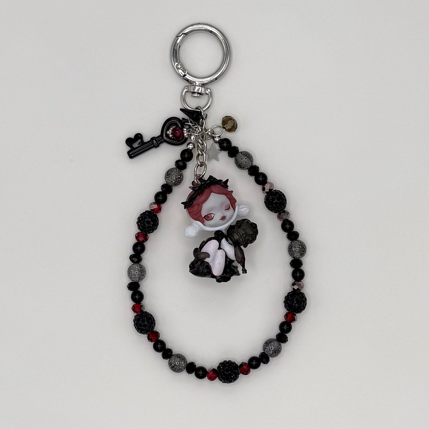 Beaded keychain with black faceted crystal, dark red crystal, and silver glitter beads with silver spacers, featuring a red-haired character figure in a black outfit hugging a dark companion figure, with a black heart key charm and smoky crystal charm on a silver ring clasp.
