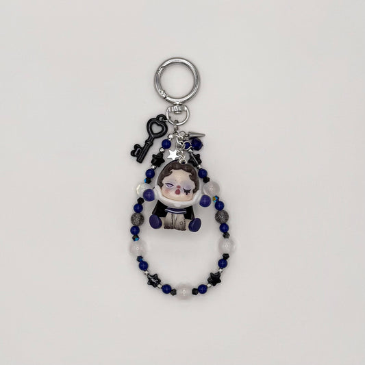 Beaded keychain with deep blue, frosted white, black star, and silver glitter beads, featuring an astronaut-helmeted girl character figure, with a black heart key charm, silver spike charm, and star charm on a silver ring clasp.